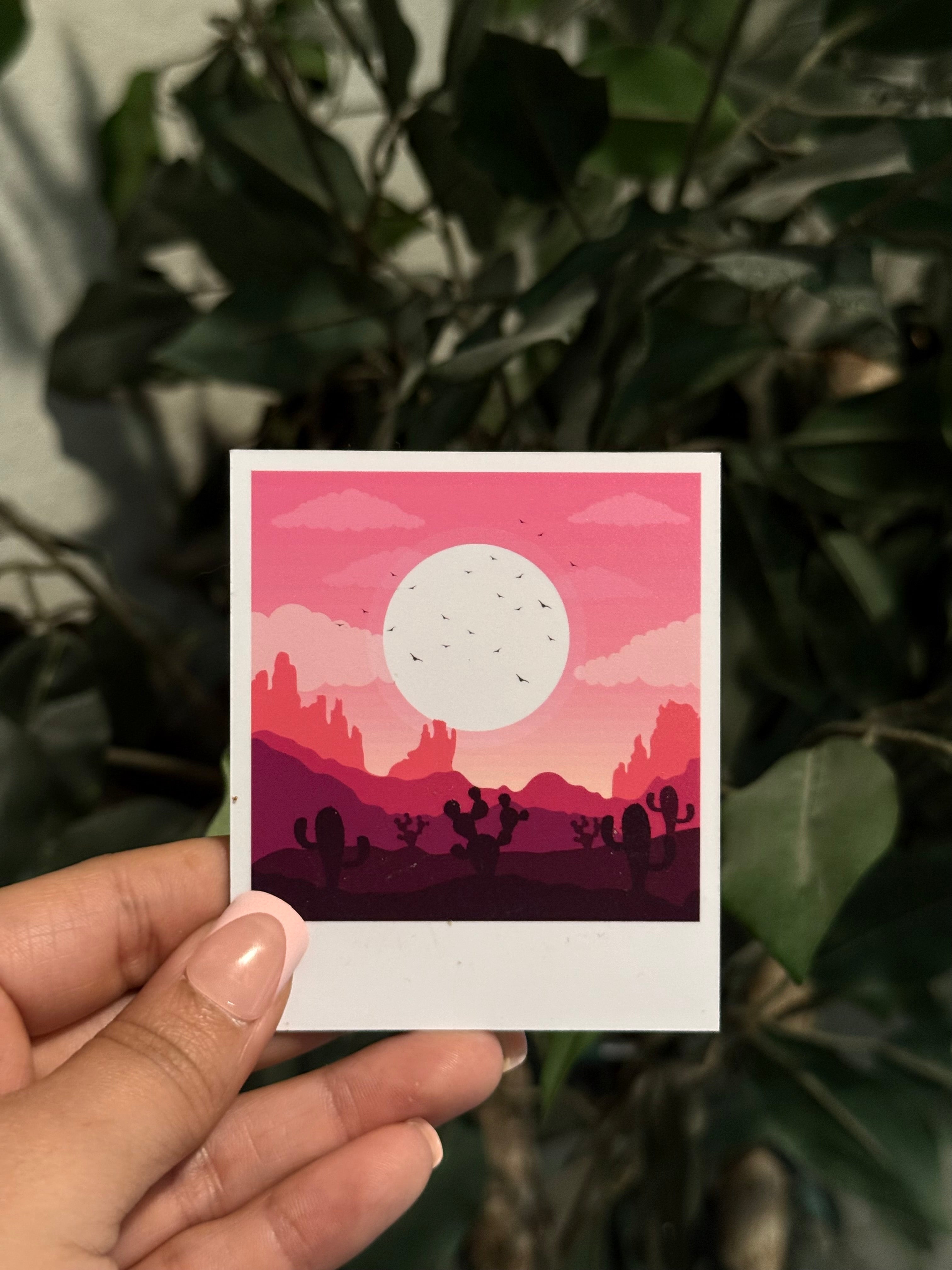 Hand holding a small polaroid personalized photo magnet with a pink and purple desert scene against a green leafy background